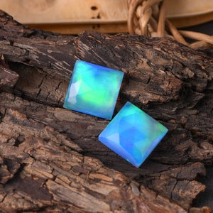 Pode incluir: Duas pedras preciosas de opala iridescente de forma quadrada, uma azul e outra verde, sobre um pedaço de madeira desgastada.