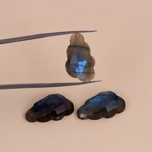 May include: Three faceted labradorite gemstones carved into cloud shapes. One stone is held by metal tweezers, displaying a dark grey base with iridescent blue flashes, while two identical stones rest below, showing similar dark tones and blue light.