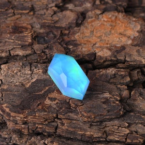 Peut inclure: Une pierre précieuse bleu clair, taillée à facettes, de forme hexagonale. La gemme est translucide, avec des nuances de vert et de bleu. Elle est posée sur une surface en bois brun texturé, créant un contraste de couleurs et de textures.