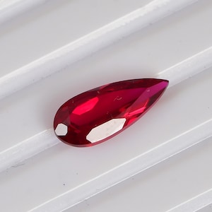 以下が含まれることがあります： 深く鮮やかな赤色をした、洋梨型のファセットカットが施された宝石。尖った先端と丸みを帯びた底部を持ち、複数の平らで磨かれた面が光を反射し、宝石全体に輝きと透明感を与えている。