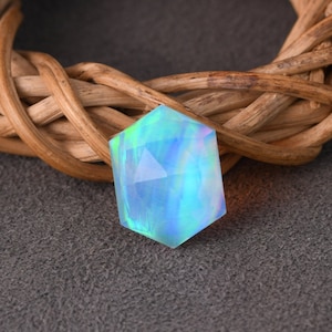 Peut inclure: Une pierre précieuse hexagonale facettée aux teintes irisées de bleu, vert et violet. La pierre est posée sur un fond de matériau tissé brun clair et une surface grise. Les facettes de la pierre captent la lumière.