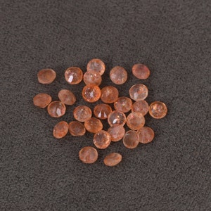 May include: A collection of 20 small, round, orange sunstone gemstones. The stones are translucent and have a shimmery, iridescent quality.