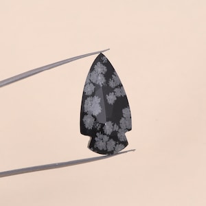 Peut inclure: Un pendentif en forme de pointe de flèche en obsidienne à flocons de neige noir et blanc. La pointe de flèche est de couleur noire foncée avec des motifs de flocons de neige blancs sur toute sa surface.