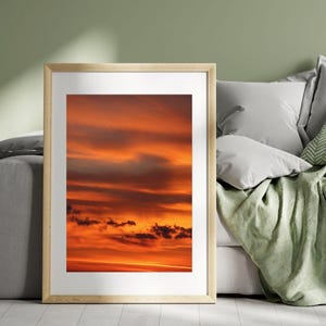 Könnte beinhalten: Ein gerahmter vertikaler Wandkunstdruck, der einen lebendigen Sonnenuntergangshimmel mit tiefen orangefarbenen, roten und goldenen Tönen zeigt. Das Bild zeigt geschichtete Wolken, die von der Sonne beleuchtet werden, in einem hellen Holzrahmen mit weißem Passepartout.