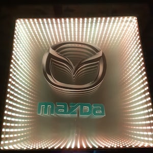 Op de afbeelding: Een 3D-geprint Mazda-logo in tealblauw met een witte omtrek, weergegeven in een gespiegelde oneindigheidsbox met witte LED-verlichting. De doos heeft een zwart frame en een wit label aan de onderkant met de tekst "Infinity Design" en "www.infinity3d.ro".