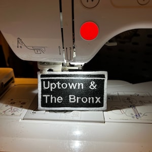 May include: A black and white embroidered patch with the text "Uptown & The Bronx" on a white background. The patch is being sewn on a white sewing machine.