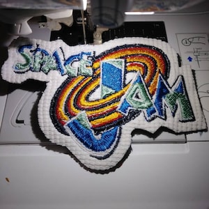 May include: A white fabric patch with colorful embroidered text that reads "Space Jam". The patch is being sewn onto a white surface with a sewing machine.