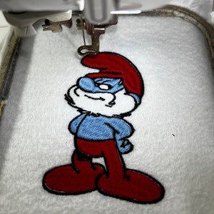 May include: A blue and red embroidered patch of a cartoon character, Smurf, with a white beard and a red hat. The patch is being sewn onto a white fabric.