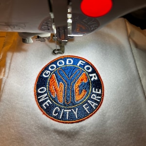 May include: Close-up of a white fabric with a circular embroidered patch. The patch is blue, orange, and yellow with the text "GOOD FOR ONE CITY FARE" and a stylized "NYC" logo.