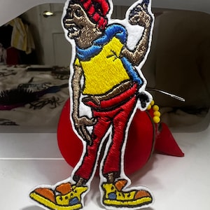 May include: Embroidered patch of a cartoon character wearing a red hat, yellow shirt, red pants, and blue shoes. The character is brown and has a white outline.