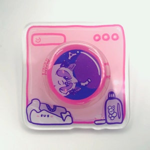 May include: A pink and white plastic washing machine with a round, purple and pink sticker featuring a cartoon raccoon inside a bubble. The sticker has a white star and a white outline. The washing machine has a white outline and the text "000" on the top.