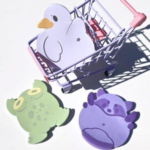 May include: A whimsical image features a miniature shopping cart in a soft purple hue, holding a white duck-shaped cutout with a yellow beak. Two additional cutouts, a green frog and a purple raccoon, are placed nearby.