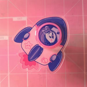 May include: A pink and blue rocket ship with a white star and the word "KOODLE" on the side. A cartoon character with black hair and a pink nose is in the window of the rocket.