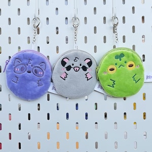 May include: Three plush coin purses with keychains. The purses are purple, gray, and green. Each purse has a different animal face embroidered on it. The purple purse has a raccoon face, the gray purse has a panda face, and the green purse has a frog face.