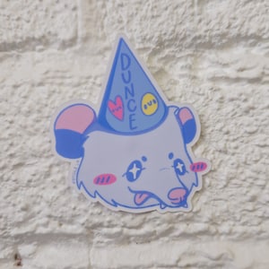 May include: A blue and white sticker of a cartoon opossum wearing a blue party hat with the word "DUNCE" written on it. The opossum has pink ears, pink cheeks, and yellow eyes.