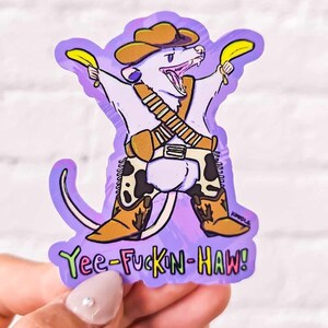 May include: A purple and white sticker featuring a cartoon possum wearing a cowboy hat and holding two bananas. The possum is sporting cowboy boots and a belt with a gun holster. The text "Yee-Fuckin-Haw!" is written beneath the possum.