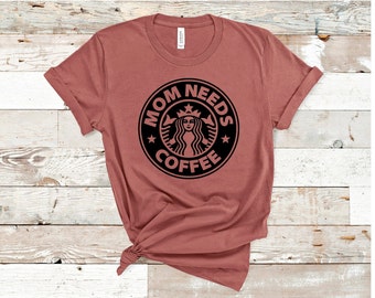 May include: A dusty rose-colored t-shirt with a black graphic. The graphic is a circular design with the words "MOM NEEDS COFFEE" around a central image of a woman. The shirt is knotted at the bottom.