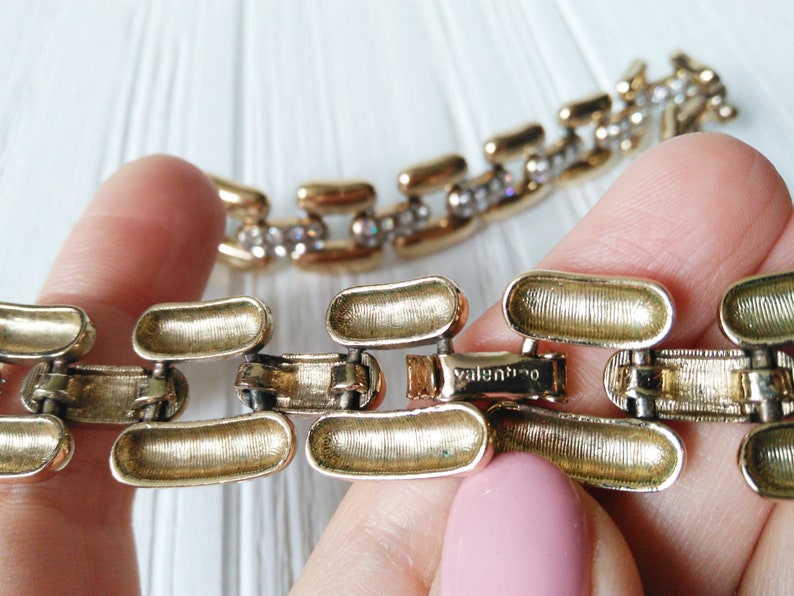 May include: Gold-toned chain link bracelet with a textured finish. The bracelet features a clasp with the brand name "Valentino" engraved. Some links are adorned with small, clear crystals, adding sparkle to the jewelry.