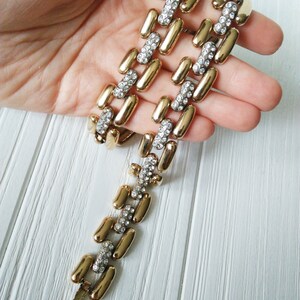 May include: Gold-tone bracelet with a series of elongated, rectangular links. Each link is connected by smaller, rectangular links encrusted with clear rhinestones. The bracelet is displayed against a white wooden surface.