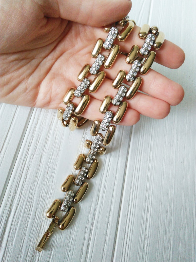 May include: Gold-tone bracelet with a series of elongated, rectangular links. Each link is connected by smaller, rectangular links encrusted with clear rhinestones. The bracelet is displayed against a white wooden surface.