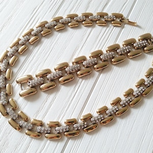 May include: A gold-toned necklace and bracelet set. The jewelry features a linked design with rectangular gold segments interspersed with sections of clear, sparkling rhinestones. The set is displayed on a white surface.
