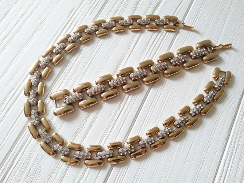 May include: A gold-toned necklace and bracelet set. The jewelry features a linked design with rectangular gold segments interspersed with sections of clear, sparkling rhinestones. The set is displayed on a white surface.
