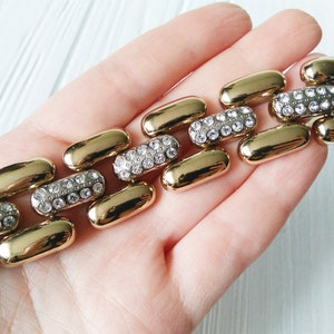 May include: Gold-toned bracelet featuring a series of elongated, pill-shaped links. Alternating links are encrusted with clear, sparkling rhinestones. The bracelet is held against a light background, showcasing its polished finish.