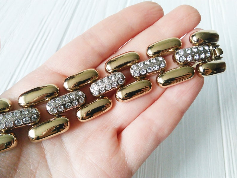 May include: Gold-toned bracelet featuring a series of elongated, pill-shaped links. Alternating links are encrusted with clear, sparkling rhinestones. The bracelet is held against a light background, showcasing its polished finish.