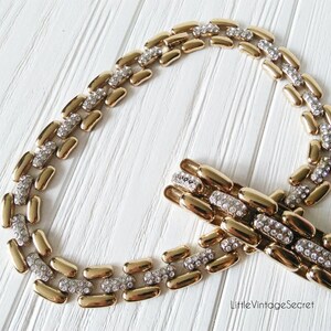 May include: A gold-toned necklace and bracelet set. The necklace and bracelet feature a linked design with alternating gold-colored and silver-colored sections. The silver sections are encrusted with small, sparkling rhinestones. The jewelry is displayed on a white wooden surface.