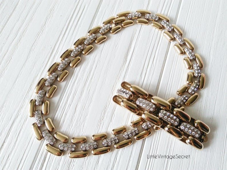 May include: A gold-toned necklace and bracelet set. The necklace and bracelet feature a linked design with alternating gold-colored and silver-colored sections. The silver sections are encrusted with small, sparkling rhinestones. The jewelry is displayed on a white wooden surface.