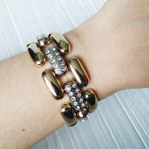 May include: Gold-toned bracelet with a series of elongated, rounded links. Alternating links are adorned with clear, sparkling crystals. The bracelet is worn on a wrist, showcasing its design and texture.