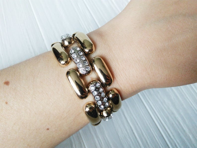 May include: Gold-toned bracelet with a series of elongated, rounded links. Alternating links are adorned with clear, sparkling crystals. The bracelet is worn on a wrist, showcasing its design and texture.