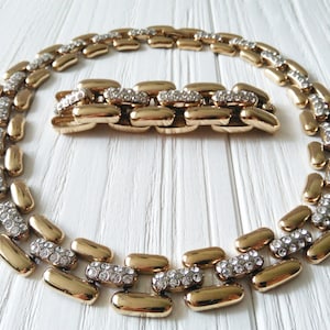 May include: A gold-toned necklace and bracelet set. The jewelry features a linked design with alternating sections of gold and sections encrusted with small, clear crystals. The pieces are displayed on a white wooden surface.