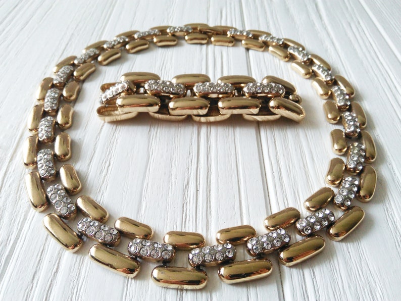 May include: A gold-toned necklace and bracelet set. The jewelry features a linked design with alternating sections of gold and sections encrusted with small, clear crystals. The pieces are displayed on a white wooden surface.