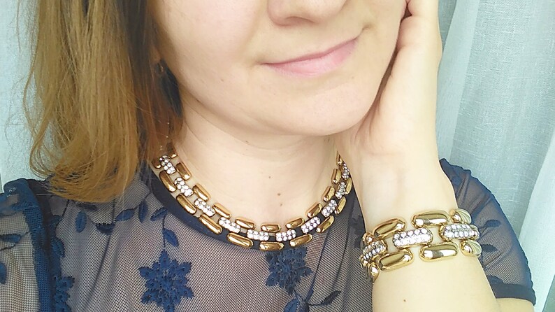 May include: Gold-toned necklace and bracelet set. The jewelry features rectangular links with a central row of clear crystals. The necklace sits around the neck, and the bracelet is on the wrist.