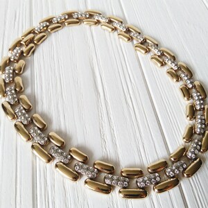 May include: A gold-toned necklace with a repeating pattern of elongated, rounded links. Each link is connected by a section of small, clear crystals. The necklace is displayed on a white wooden surface.