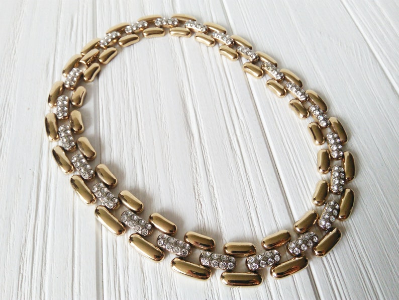 May include: A gold-toned necklace with a repeating pattern of elongated, rounded links. Each link is connected by a section of small, clear crystals. The necklace is displayed on a white wooden surface.