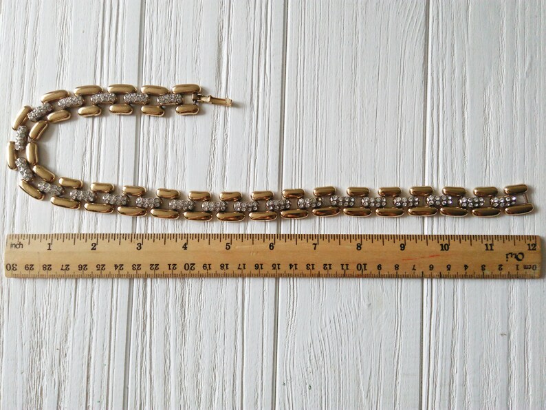 May include: A gold-toned necklace with rectangular links, each accented with a row of small, clear rhinestones. The necklace is approximately 12 inches long, as measured by a wooden ruler.