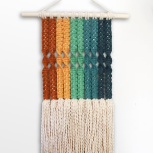 Könnte beinhalten: Ein Wandbehang aus Makramee mit einem Regenbogen aus Farben, darunter Orange, Gelb, Grün und Blau. Der Behang hat unten einen weißen Fransen.