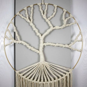 Könnte beinhalten: Ein weißer Makramee-Wandbehang in Form eines Baumes mit einem goldenen Metallring. Der Baum hat einen dicken Stamm und Äste, die sich kreisförmig ausbreiten.