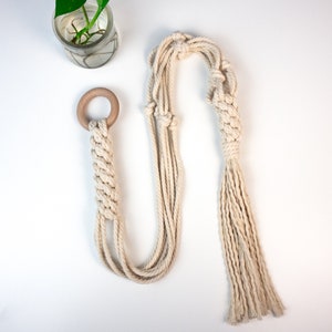 May include: A macrame plant hanger with a wooden ring, made from natural cotton rope. The hanger has a long tassel and is designed to hang from the ceiling.