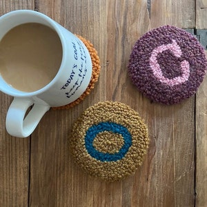 May include: A white mug with a beverage rests on an orange, circular coaster. Two additional coasters are visible: one with a purple background and a pink "C", and another with a gold background and a teal circle. The coasters are on a wooden surface.