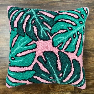 Puede incluir: Una almohada de tiro rosa y verde con un patrón de hojas de monstera. La almohada está hecha de una tela suave y mullida y es perfecta para agregar un toque de estilo tropical a cualquier habitación.