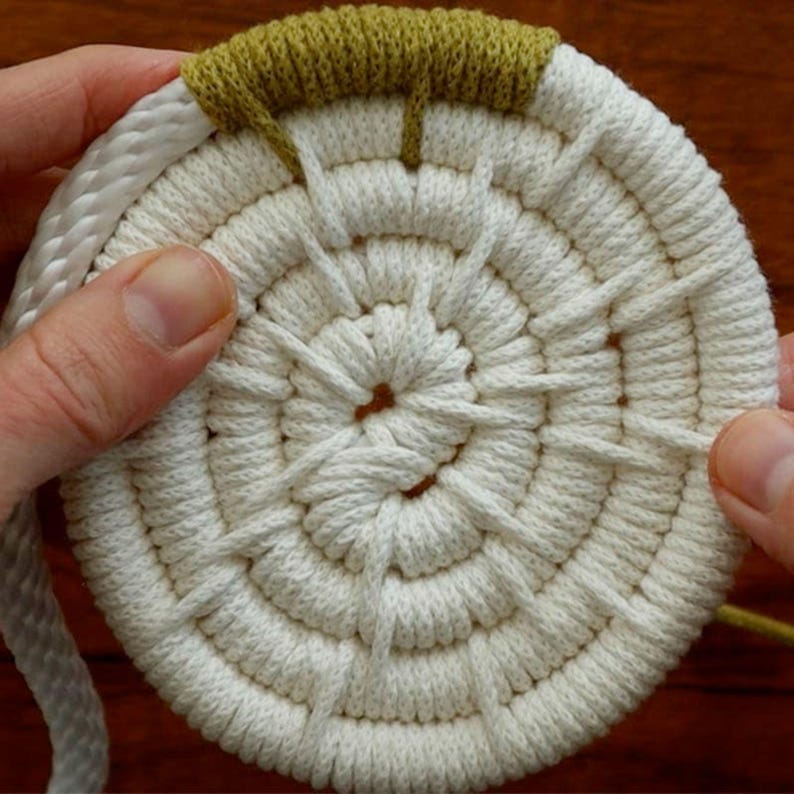 May include: A handmade, circular coaster crafted from off-white rope, with a central design and a green accent on the edge. The coaster is held by two hands, showcasing the texture and construction of the item.