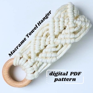 May include: Cream-coloured macrame towel hanger with a wooden ring. The text "Macrame Towel Hanger" and "digital PDF pattern" are visible. The macrame is intricately knotted, creating a decorative and functional design.