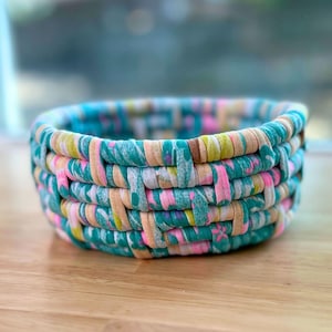 May include: A round woven basket constructed from strips of fabric featuring a multicolored pattern of teal, pink, yellow, and white. The basket has a thick, textured rim and a repeating interlaced design throughout its circular structure.
