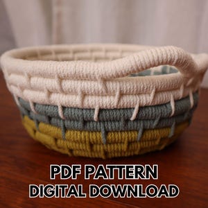 Puede incluir: Una cesta redonda hecha a mano con un diseño tejido. La cesta presenta una parte superior color crema, una sección media gris-verdosa y una base amarillo-verdosa. Tiene un asa color crema. El texto "PDF PATTERN DIGITAL DOWNLOAD" está en la parte inferior.
