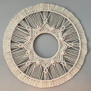 Op de afbeelding: Een grote, ronde macramé wandhanger met een wit, ingewikkeld ontwerp. De hanger heeft een centrale cirkel met een stervormig patroon en een franje rand.