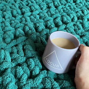 May include: A light purple ceramic mug with the word "FOCUS" printed on the side. The mug is filled with coffee and is sitting on a green fuzzy surface.