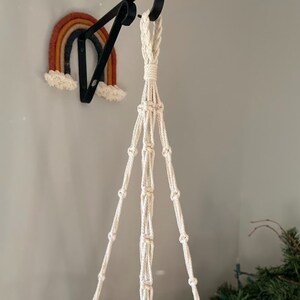 May include: A cream-colored macrame plant hanger suspended from a black metal hook. The plant hanger is knotted with a series of decorative knots. A felt rainbow in shades of orange, yellow, and blue is mounted on the wall.